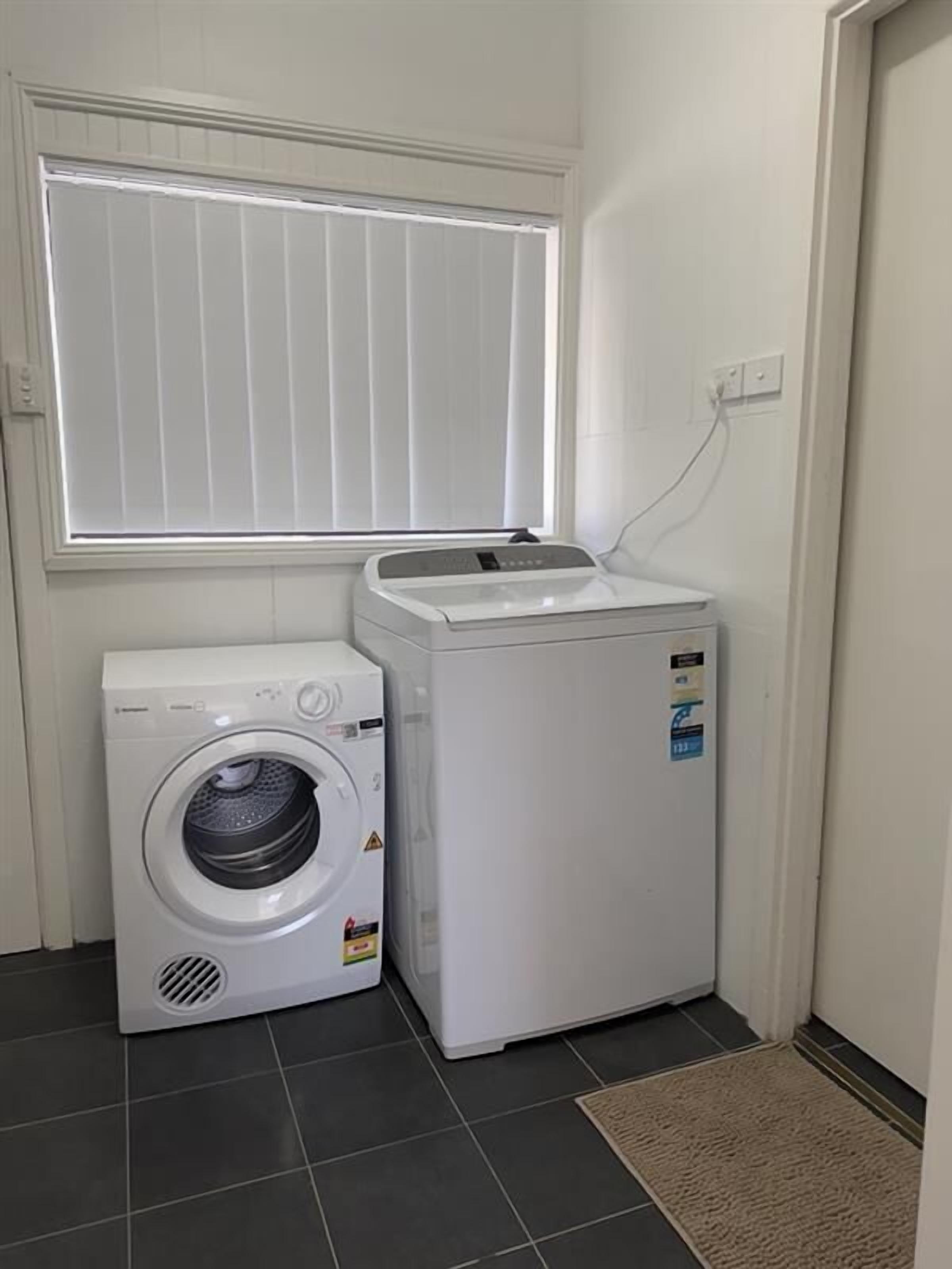 Un espace buanderie comprenant un lave-linge à chargement frontal et un sèche-linge, une fenêtre avec des stores et un sol carrelé.