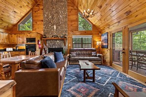 A log cabin interior with a stone fireplace, wooden furniture, and a large window overlooking the outdoors. - Cavalier Cabin - Pet Friendly | Hot Tub | Mountain View | Game Room (Blue Ridge)