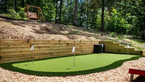 Une aire de jeux avec une balançoire en bois, une zone recouverte de gazon synthétique vert et ornée de drapeaux, et un banc en bois.