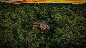 A two-story cabin surrounded by dense forest, with a visible deck and outdoor seating area. - Cavalier Cabin - Pet Friendly | Hot Tub | Mountain View | Game Room (Blue Ridge)