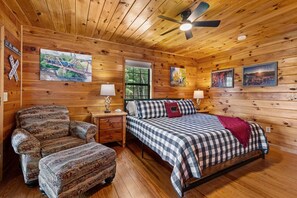 A wooden cabin interior with a bed, a chair, a nightstand, and a ceiling fan. - Cavalier Cabin - Pet Friendly | Hot Tub | Mountain View | Game Room (Blue Ridge)