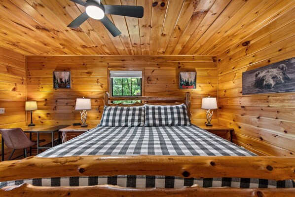 A log cabin bedroom with a bed, two bedside tables, two lamps, a ceiling fan, and framed pictures on the walls.