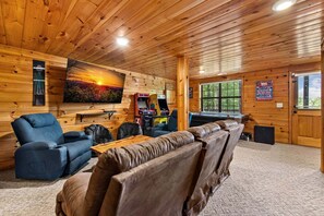 A cozy cabin interior with a large flat-screen TV, gaming consoles, a leather couch, and a window overlooking greenery.