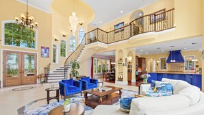 Amplia sala de estar con una escalera, muebles blancos y azules, y una cocina al fondo.