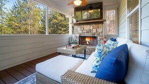Una habitación acogedora con chimenea de piedra, ventilador de techo y un gran ventanal con vistas a árboles.