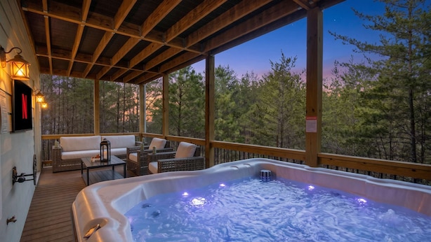Una terraza de madera con un jacuzzi, mobiliario de mimbre y vistas a árboles.