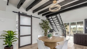 Un comedor moderno con una mesa de madera y sillas blancas, una planta grande en maceta y una escalera que lleva a un patio exterior.