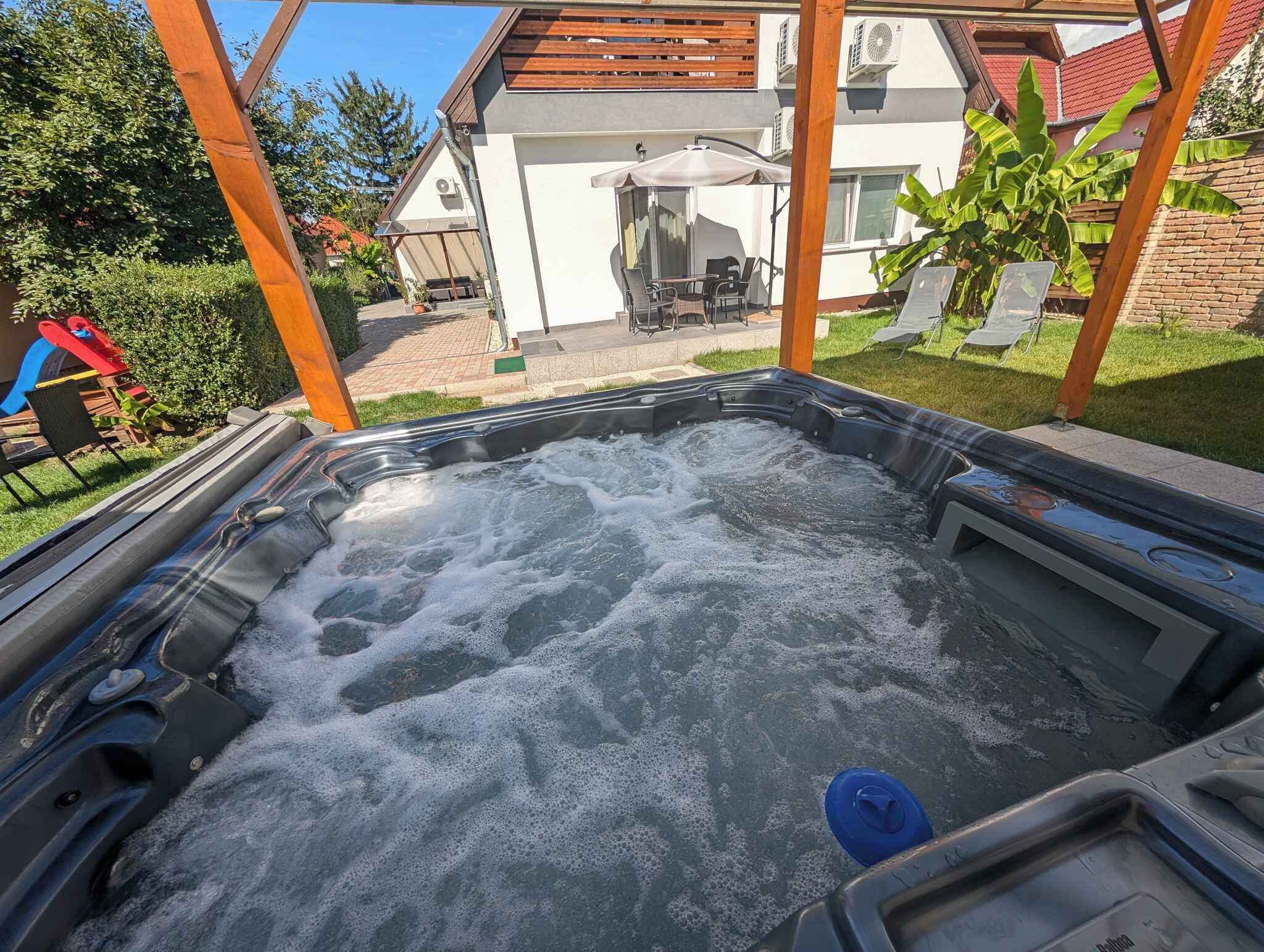 A hot tub with a wooden pergola overhead, surrounded by a backyard with a patio, outdoor furniture, and a house in the background.