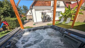 A hot tub with a wooden pergola overhead, surrounded by a backyard with a patio, outdoor furniture, and a house in the background.