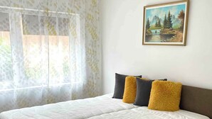 A bedroom with a large bed, two pillows, a painting on the wall, and a window with sheer curtains.