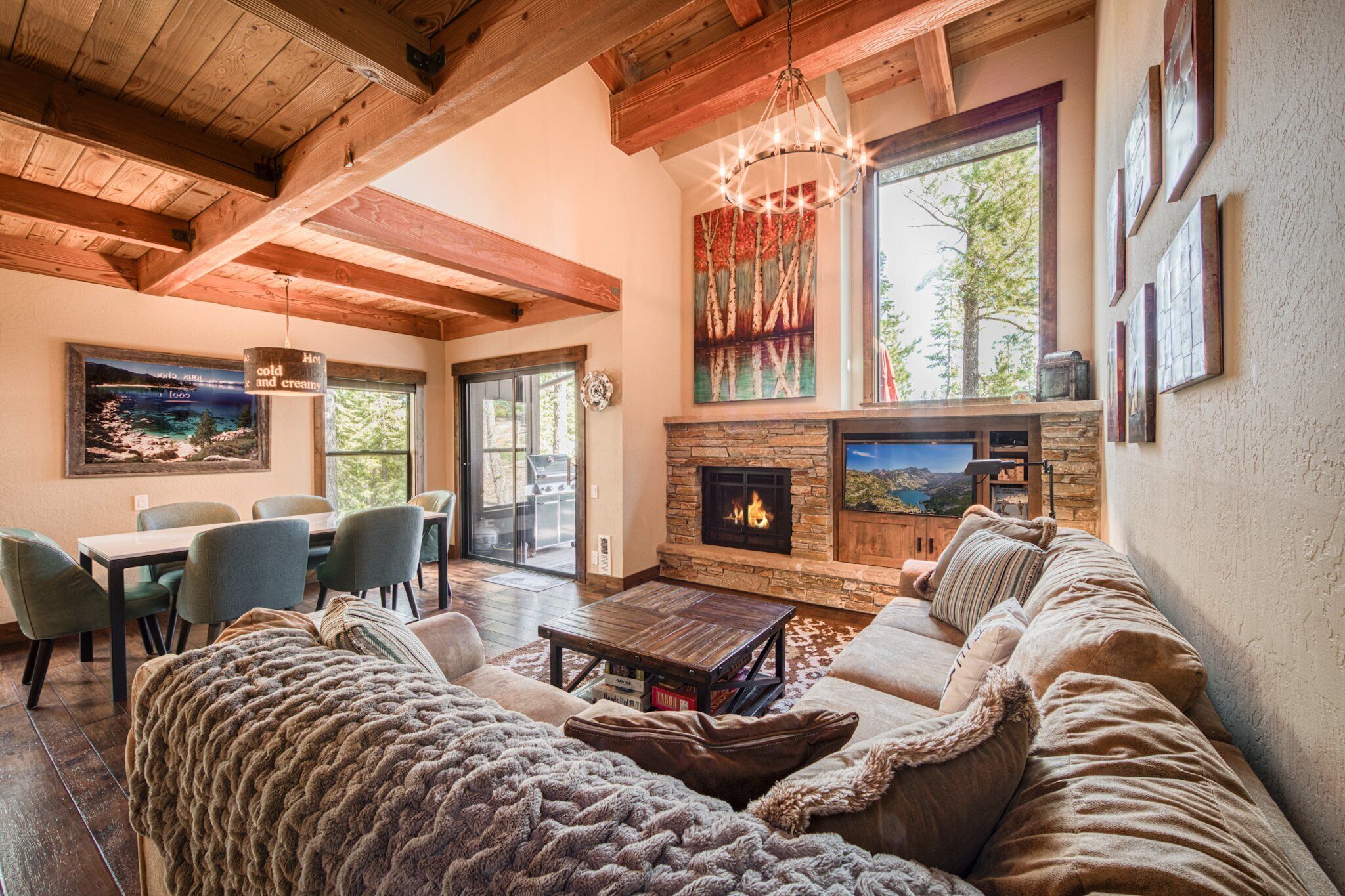A cozy living room with a fireplace, wooden beams, a dining area, and large windows.