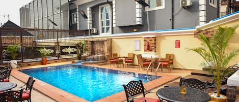 Una piscina con sillas de descanso y mesas, un patio techado y un edificio con fachada gris y ventanas amplias.