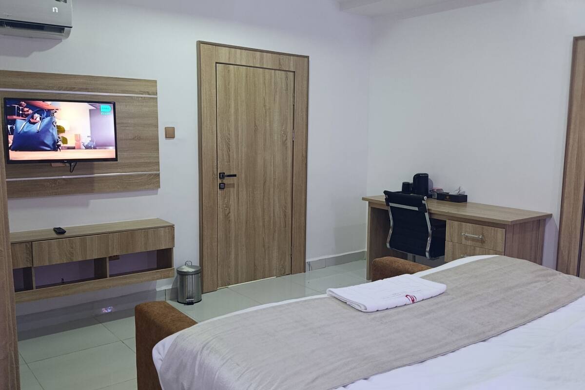 A hotel room with a bed, a TV mounted on the wall, a desk with a chair, and a wardrobe.