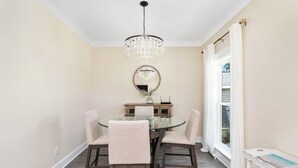 Un coin repas comprenant une table ronde en verre, quatre chaises, un lustre, un miroir et une fenêtre avec des rideaux.