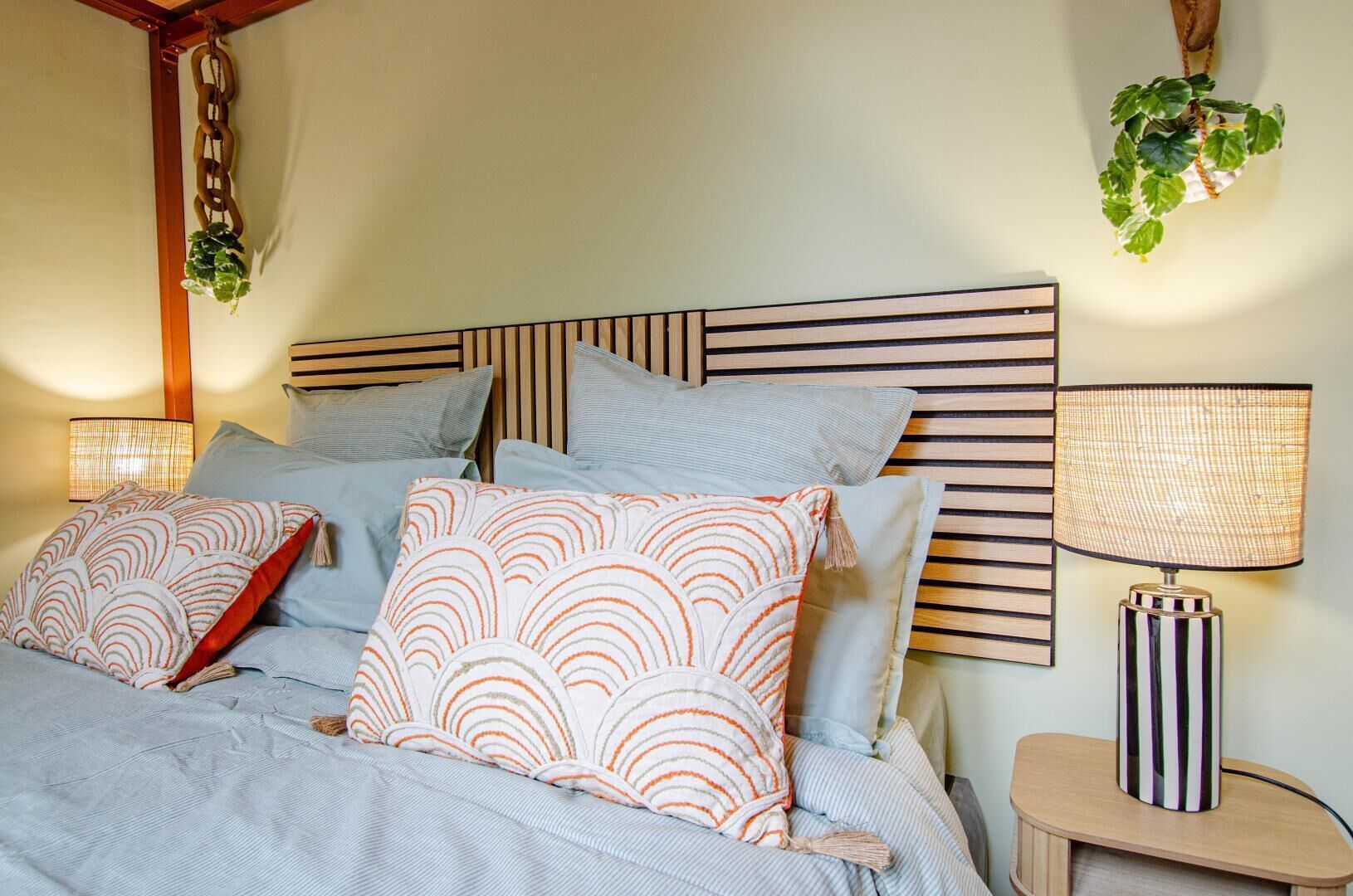 A neatly made bed with striped bedding and patterned pillows, a bedside lamp, and a hanging plant.