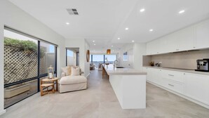 A modern kitchen with a central island, white cabinetry, and a view of the outdoors through large windows. - Three-Bedroom Luxurious Beachfront Holiday Home (North Brighton)