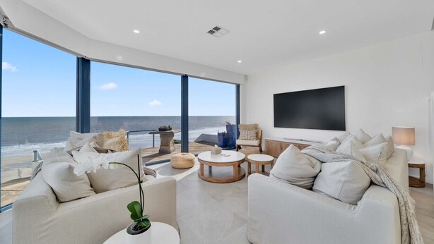A modern living room with a large window overlooking the ocean, a flat-screen TV mounted on the wall, and a cozy seating area with a coffee table. - Three-Bedroom Luxurious Beachfront Holiday Home (North Brighton)