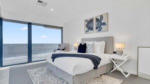 A modern bedroom with a large bed, a nightstand, and a view of the ocean. - Three-Bedroom Luxurious Beachfront Holiday Home (North Brighton)