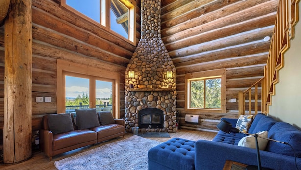 L’intérieur d’une cabane en rondins, avec une cheminée en pierre, des planchers en bois, un canapé en cuir marron, un canapé bleu et de grandes fenêtres.