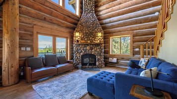 L’intérieur d’une cabane en rondins, avec une cheminée en pierre, des planchers en bois, un canapé en cuir marron, un canapé bleu et de grandes fenêtres.