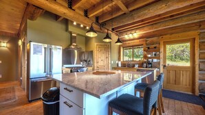 A log cabin kitchen with a stainless steel refrigerator, a large island with bar stools, wooden beams on the ceiling, and a window overlooking greenery.