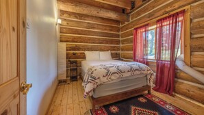 A log cabin room with a bed, wooden walls, a window with red curtains, and a patterned rug.