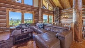 L’intérieur d’une cabane en rondins, avec un grand canapé d’angle en cuir, des poutres en bois et de grandes fenêtres offrant une vue sur l’extérieur.