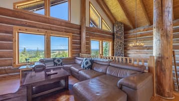 L’intérieur d’une cabane en rondins, avec un grand canapé d’angle en cuir, des poutres en bois et de grandes fenêtres offrant une vue sur l’extérieur.