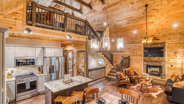 A spacious log cabin interior with a kitchen, dining area, and living room, featuring wooden floors, a fireplace, and a staircase. - Modern Rustic 3BR & Loft, Hot Tub, GR and Fire Pit (Ellijay)