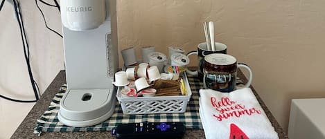 Une machine à café, un panier de dosettes de café, une tasse, une télécommande et une serviette ornée d’un message sur le thème de l’été, le tout posé sur un plan de travail de cuisine.