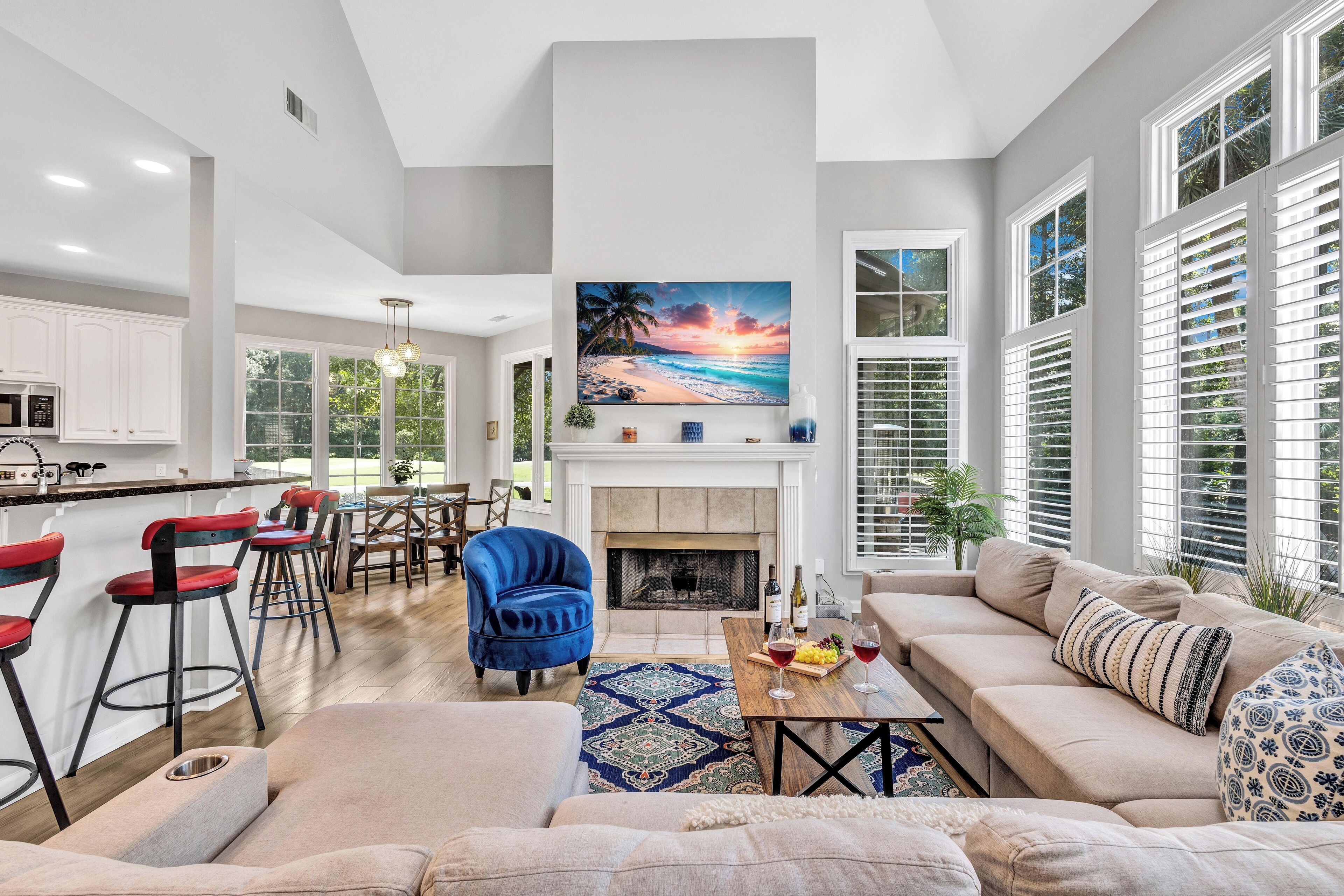A spacious living area with a fireplace, a large painting of a sunset, and a kitchen in the background.