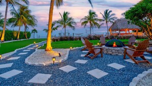 A tropical outdoor seating area with a fire pit, lounge chairs, and a thatched-roof structure.