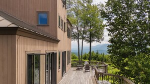 Une terrasse en bois avec une table de pique-nique et des chaises, offrant une vue sur une zone boisée et, au loin, sur des montagnes.