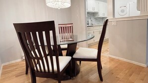 Un comedor con mesa de vidrio y sillas de madera oscura, un candelabro y una cocina al fondo.