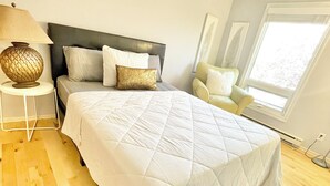 Un dormitorio con cama, mesita de noche, lámpara, silla y ventana.