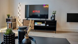 Una sala de estar moderna con un televisor de pantalla plana mostrando 'NETFLIX', una mesa de centro negra con objetos decorativos, un mueble de madera y un estilo decorativo minimalista.