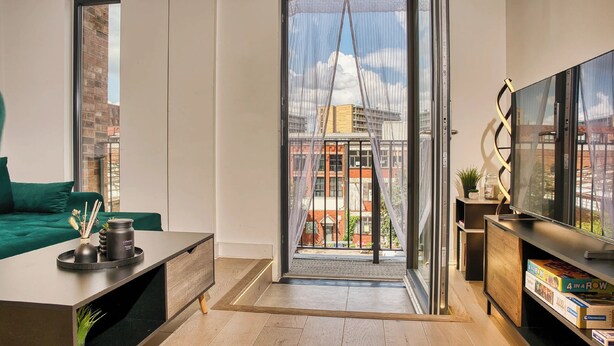 Una sala de estar moderna con piso de madera, un sofá verde, una mesa de centro con una vela y un florero, un mueble de televisión con libros y vistas a edificios a través de una puerta de vidrio.