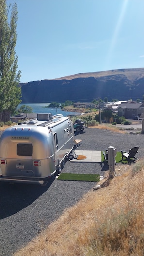 Een caravan geparkeerd op een grindpad, met een meer en bergen op de achtergrond.