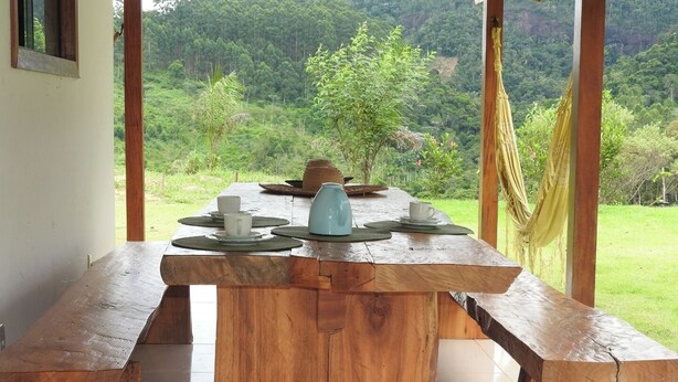 A wooden dining table set with plates, cups, and a kettle, overlooking a lush green landscape. - Sítio Martinelli, your connection with Nature, come and have a well-deserved rest. (São Lourenço Santa Teresa)