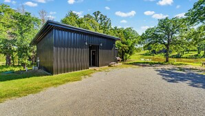 A metal building with a door, surrounded by a gravel driveway, trees, and a grassy area. - The Red Bird | Treehouse Stay + Pickleball Court (Alvord)