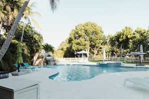 Una piscina con sillas de descanso y sombrillas, rodeada de árboles y una cerca blanca.