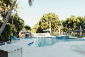 Una piscina con sillas de descanso y sombrillas, rodeada de árboles y una cerca blanca.