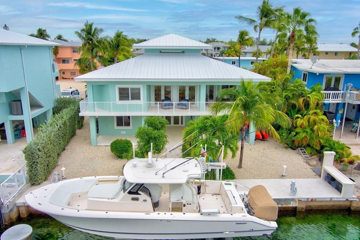 A boat docked in front of a light blue house with a balcony, surrounded by palm trees and other buildings.