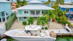 A boat docked in front of a light blue house with a balcony, surrounded by palm trees and other buildings.