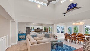 A spacious living area with a large beige sectional sofa, a dining table with wooden chairs, and a kitchen in the background.