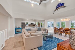 Uma sala de estar espaçosa com um amplo sofá modular bege, uma mesa de jantar com cadeiras de madeira e uma cozinha ao fundo.