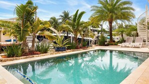 A swimming pool with lounge chairs, palm trees, and boats in the background.