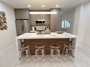 Cozinha moderna com ilha central, eletrodomésticos de aço inoxidável e banquetas de vidro transparente.