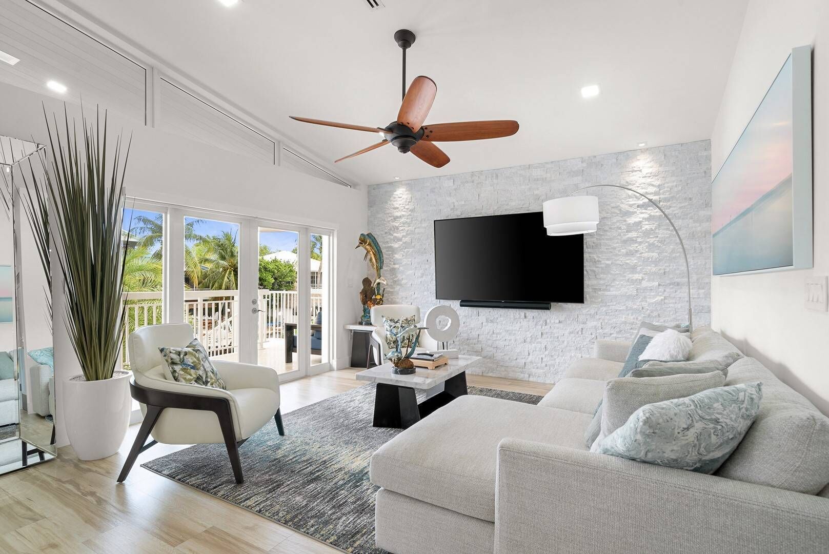 A modern living room with a ceiling fan, a flat-screen TV mounted on a textured wall, a large potted plant, and a view of the outdoors through large sliding doors.