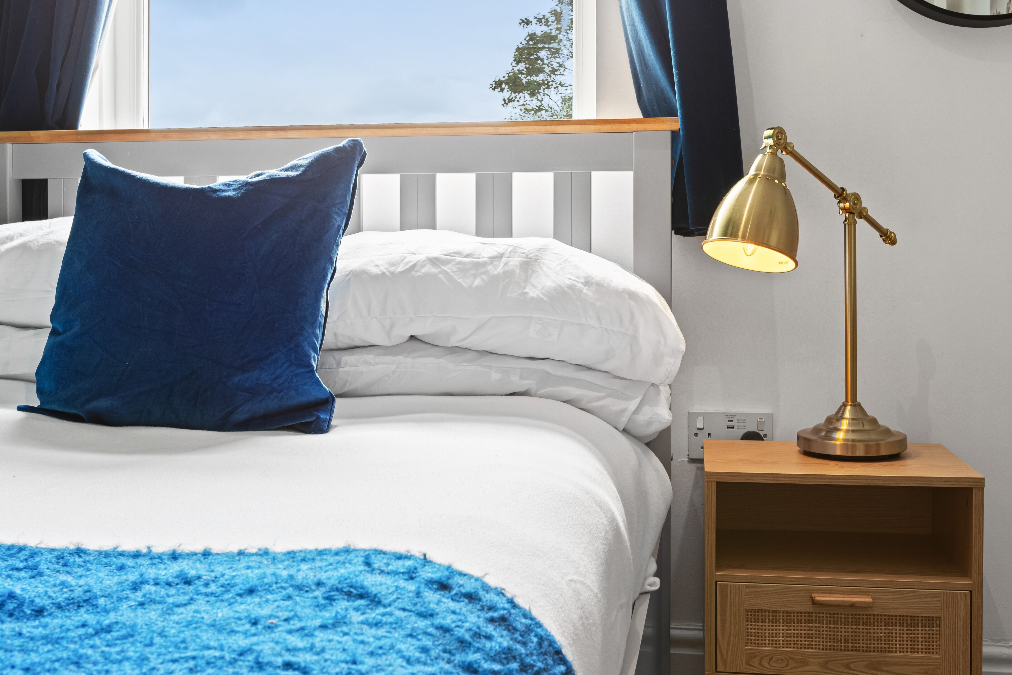 Un letto rifatto con biancheria da letto bianca e azzurra, una coperta azzurra e una lampada da comodino su un comodino in legno.